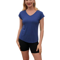 Woman wearing a blue shirt and black shorts standing in a room with a dark wall and wooden floor.