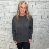 Woman wearing a gray sweatshirt and black leggings standing against a light gray brick wall.