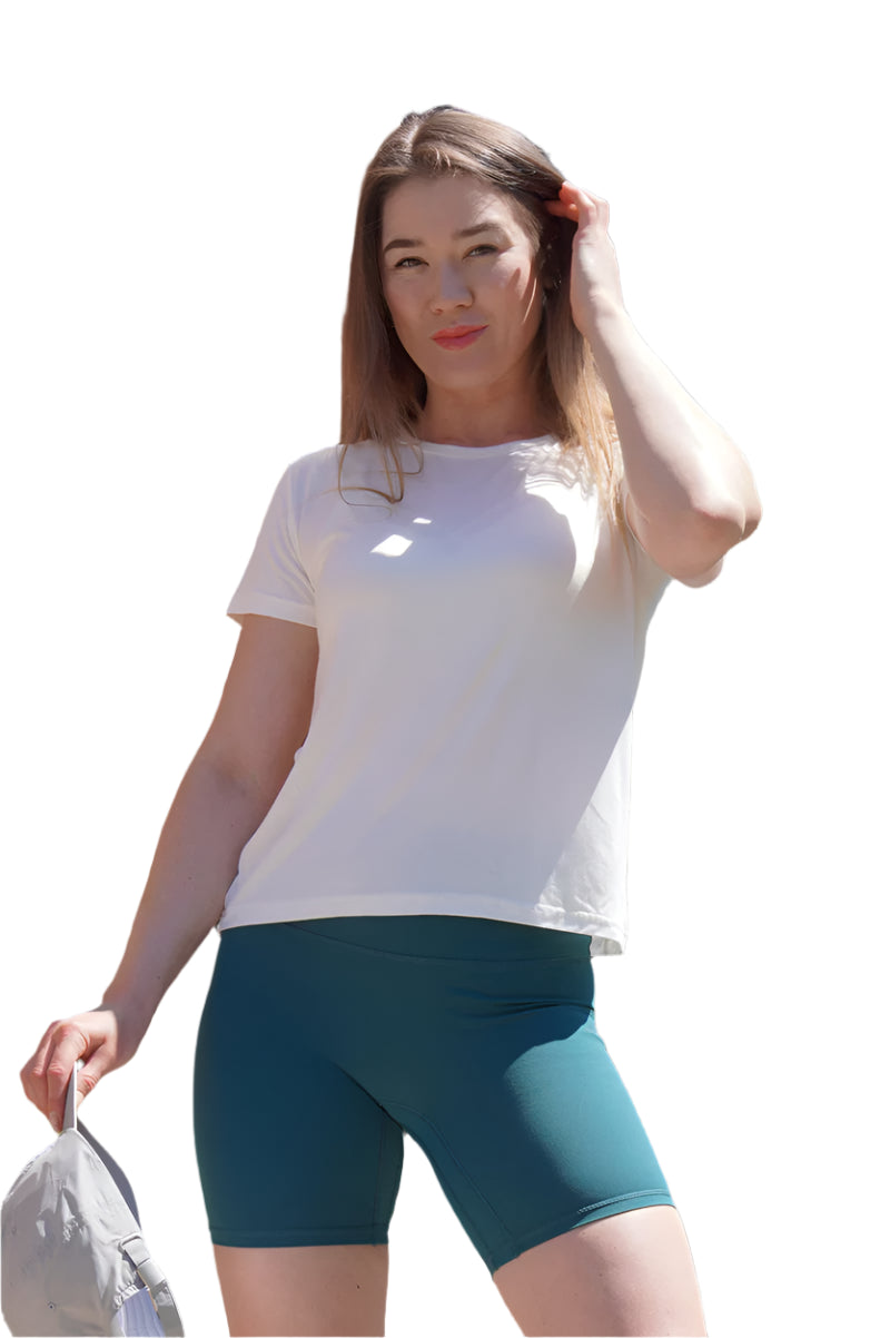 Woman wearing a white t-shirt and teal shorts standing outdoors with trees and a clear sky in the background.