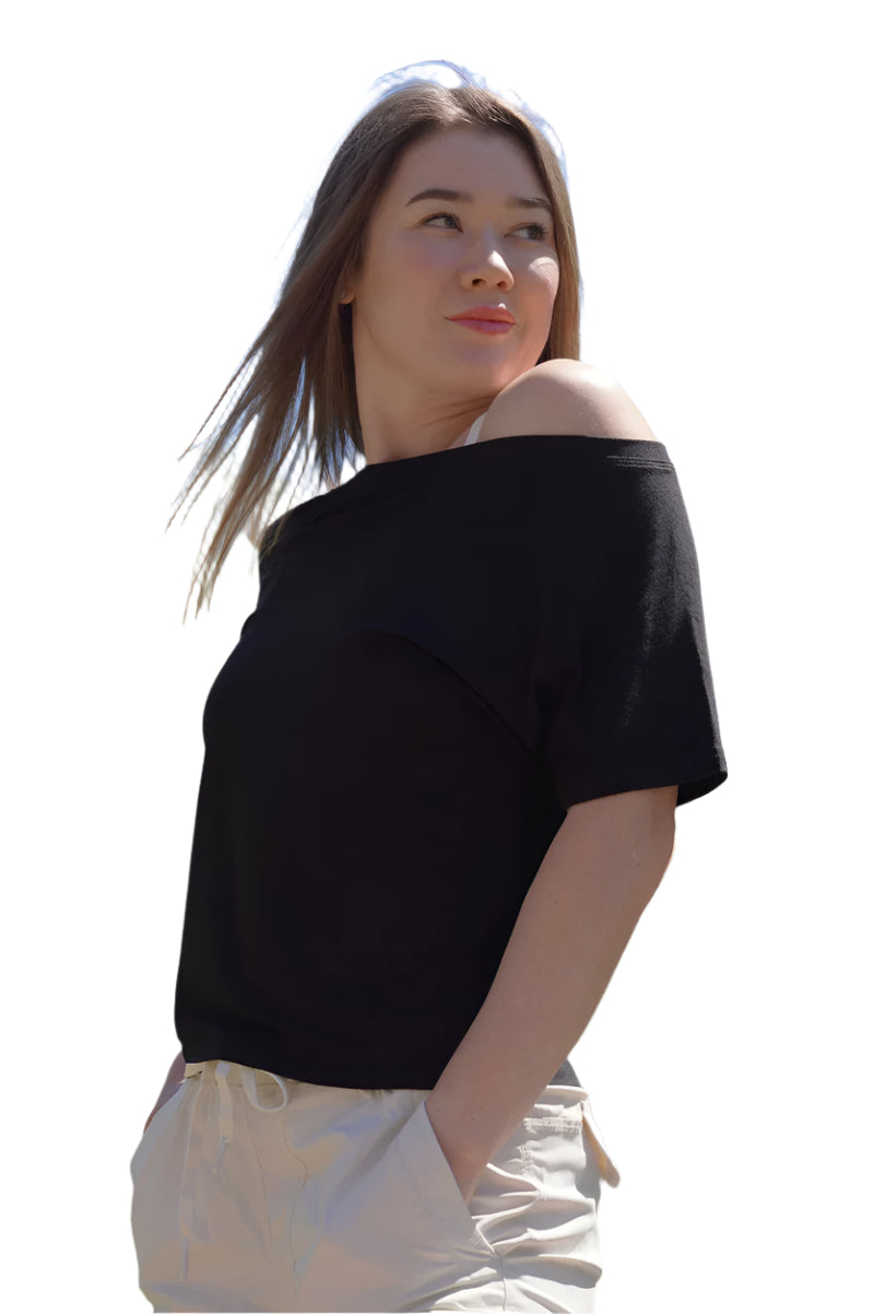Woman wearing a black off-shoulder top and light-colored pants outdoors with trees and blue sky in the background.