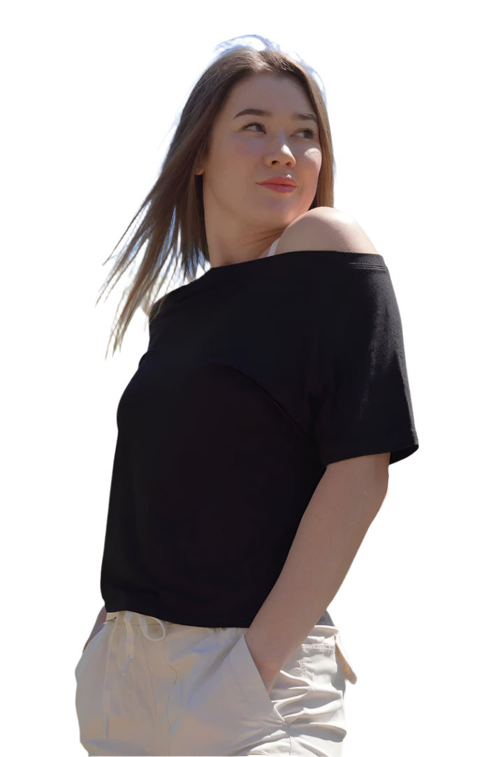 Woman wearing a black off-shoulder top and light-colored pants outdoors with trees and blue sky in the background.