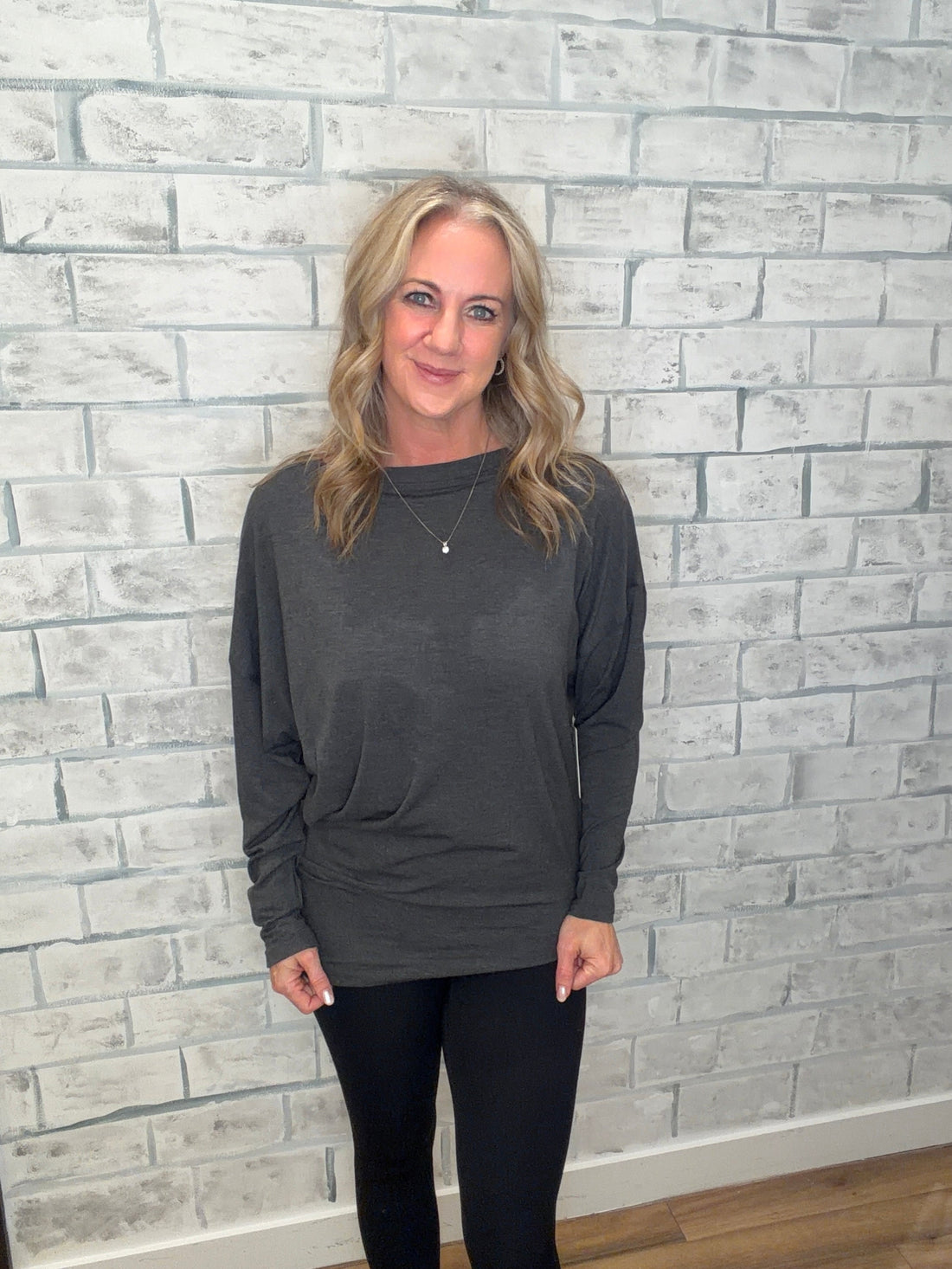 Woman wearing a gray sweatshirt and black leggings standing against a light gray brick wall.